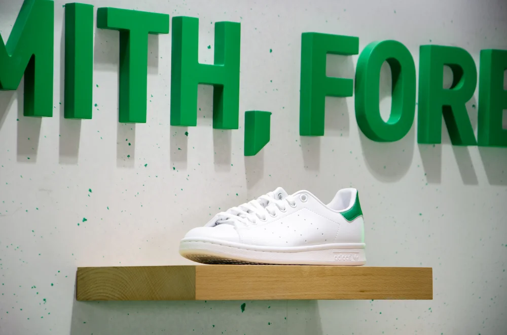 Sneaker Adidas Stan Smith exposée sur un socle bois avec un panneau en plastique recyclé pour le lancement de la Clean Classique.